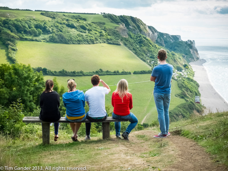 4 seated people and one standing, backs to the camera, with a view overlooking Branscombe bay, Dorset