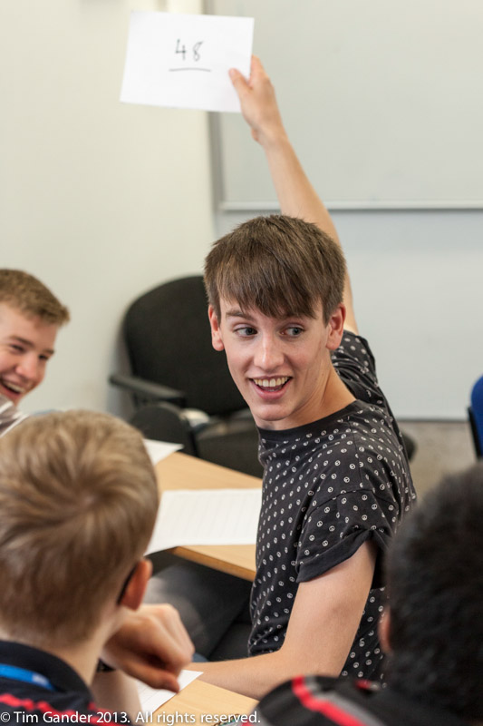 A young man in a lecture theatre holds up a white card with the number 46 written on it as part of a maths Summer camp event at University of Bath