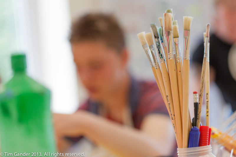 An out of focus boy sitting at a table with in-focus paint brushes in the foreground, taken for Cornerstones Schools, Warrington