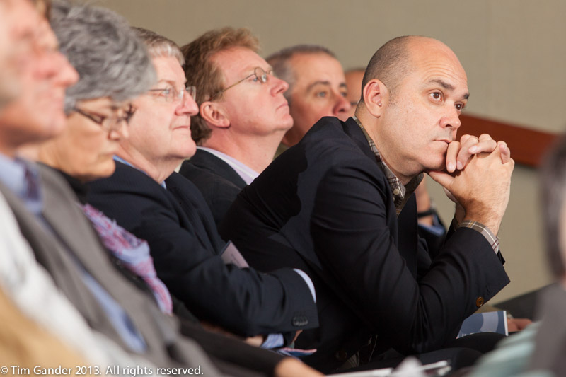 A group of seated business people in an auditorium listen to a presentation as one man leans forward to hear better