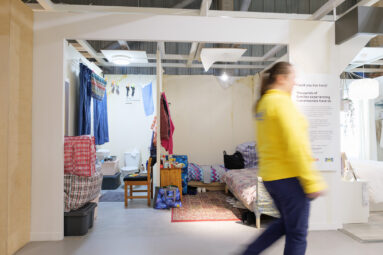 An Ikea colleague in a yellow top is blurred as they walk past a room set designed to mimic the living conditions of someone living in temporary accommodation.