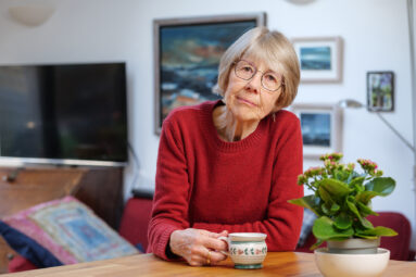 A retired woman with straight bobbed hair, in red jumper and wearing glasses leans on her kitchen counter, looking to camera with her hand on a tea mug handle.