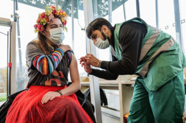 A young woman with flowery head dress for the Winter Solstice is vaccinated against Covid 19 by a pharmacist in the visitor centre cafe.