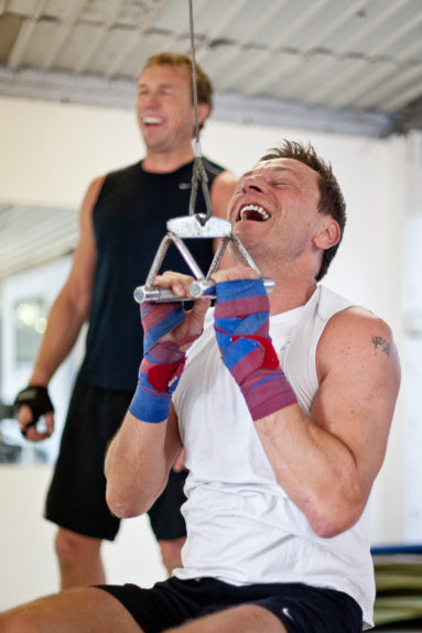 A business executive wearing a sports vest at a gym pulls weights and laughs, watched by his trainer who is also laughing.