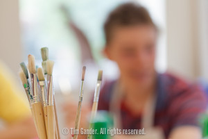 Photo focusing on a cluster of paint brushes in a pot with a boy out of focus in the background