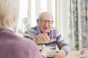 An elderly gentleman smiles and shares tea and cake with a friend who sits with their back to the camera.