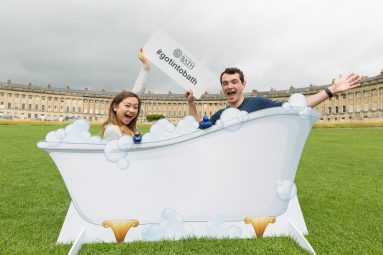 Got Into Bath social media campaign photo shows students Josh Bee and Tanya Savage posing with a cutout of a bath tub in front of Bath's Royal Crescent, holding a #gotintobath sign and waving, smiling to camera.