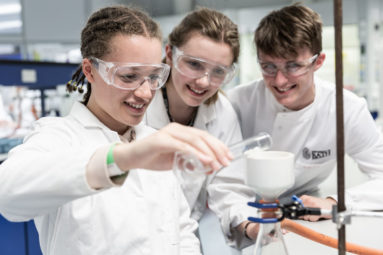 Illustrative photo of three young students in white laboratory coats, glasses and gloves working in a chemistry lab.