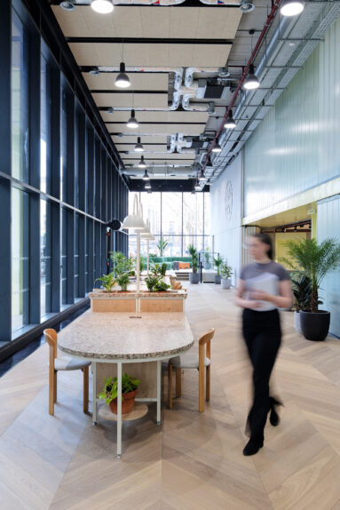 A blurred female figure walks through an open-plan office reception area with wooden floor, marble desks and glass and steel window wall.