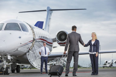 A suited business man pulling a wheeled suitcase shakes hands with a female airline executive with a jet aircraft parked beyond, the captain waiting at the foot of the steps.