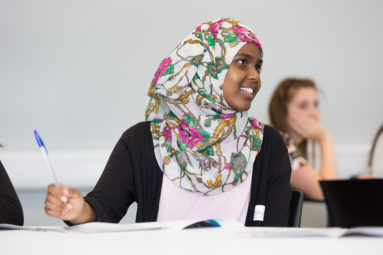 A young female student in headscarf, seated at a desk in a lecture room, smiles to someone off camera.