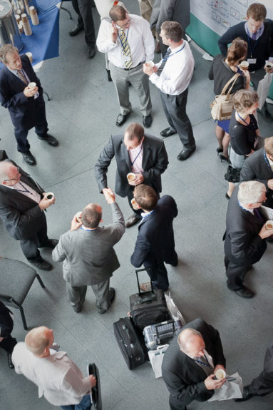 Photo taken from a high viewpoint showing a foyer of businesspeople networking over coffee with two figures shaking hands at the centre of the image.