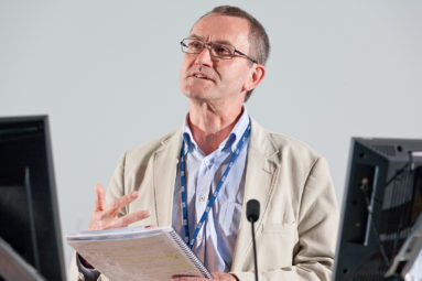 A male speaker in light fawn jacket and blue shirt speaks from notes at a University of Bath seminar.