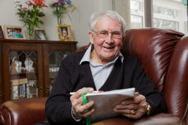 Reverend Claude Boots poses in his leather armchair at home, pen and pad in hand smiling to camera.