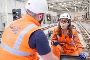 A female railway engineer in high-visibility clothing and hard hat looks towards a male colleague (his back to camera) dressed the same as they discuss technical issues inside the Bristol Parkway training shed which has all the tracks, lights and points systems of a real track.