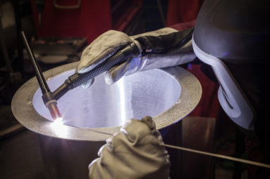 A welding engineer uses welding torch and flux rod to weld a steel collar to a tubular section of pipe.