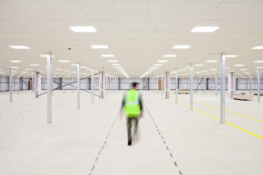 A blurred figure in high-visibility vest walks away from the camera in a large, empty, new warehouse.