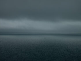 Three yachts in the far distance on flat, grey water with a moody, grey sky above. Taken from near Start Bay, South Devon.