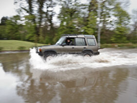 A range Rover is driven at speed through flood water.