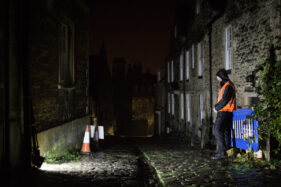 A TV security man stands in the dark of Gentle Street, Frome, at night illuminated by a floodlight.