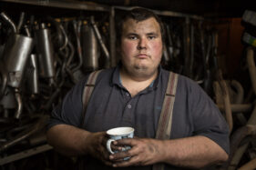 Donald of H&B Tyres in Frome, Somerset, UK, looks to camera with a cup of tea in his hand and car exhaust pipes ranged behind him. He's wearing a blue polo shirt and braces.