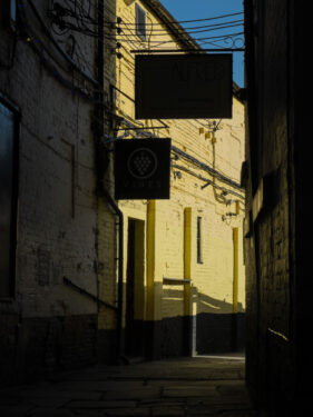 Sunlight catches a bright yellow-painted brick wall in an alley in Derby town centre, with bar signs silhouetted above.