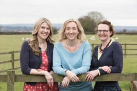 Helen Rimmer, Debbie Clifford and Michelle Gordon-Coles of Tea for Three Marketing and Communications together, looking over a wooden field gate to camera with a field of sheep behind.