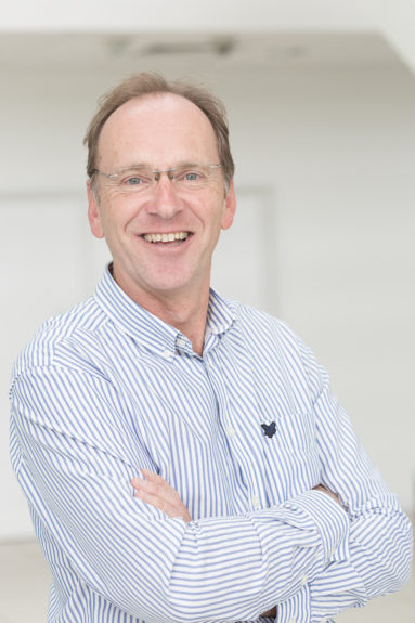 Business man in open-necked striped shirt smiles to camera.