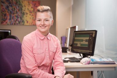 A young, stylish woman in peach blouse, hair close-cropped at the sides and swept back on top, sits side-on to her desk, smiling to camera for a business portrait.