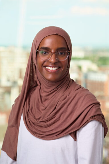 A young woman in mid-brown head scarf, white blouse and glasses smiles to camera with an out-of-focus view of a city behind her.