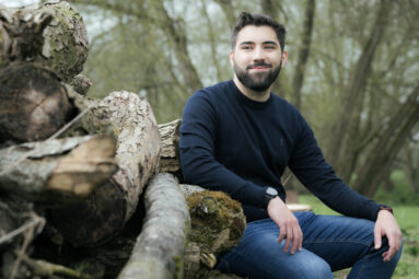 Bearded man seated besides a pile of logs.