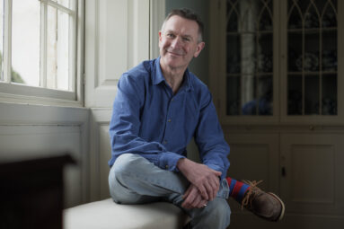 Portrait of a man in blue shirt and pale jeans sitting in a window seat of a wooden-panelled Georgian bay window, one leg up on the knee of the other with his hands resting on the knee while smiling towards the camera.