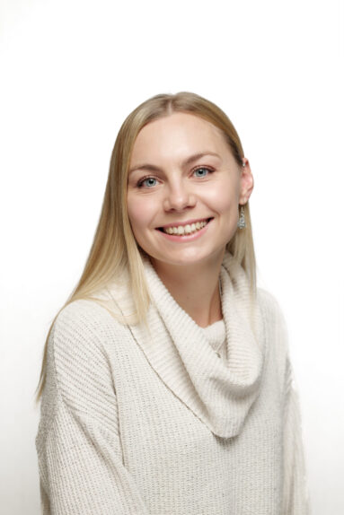 A blonde, straight haired young female business person smiles to camera. She is wearing a knitted sweater with a large draped collar and the background is white.