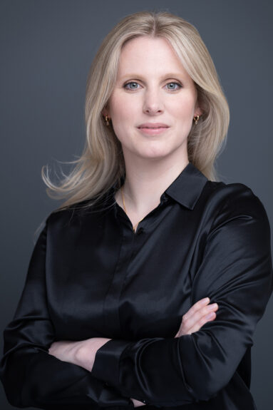 Blonde female dressed in a smart satin black blouse, smiling to camera for a business portrait.