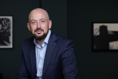 Bearded man in business jacket and shirt looking to camera with a dark green office wall behind with two framed prints on the wall which are beyond the focus of the camera.