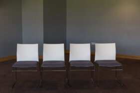 A row of 4 empty conference chairs against a grey wall with staggered corner and on brown carpet.