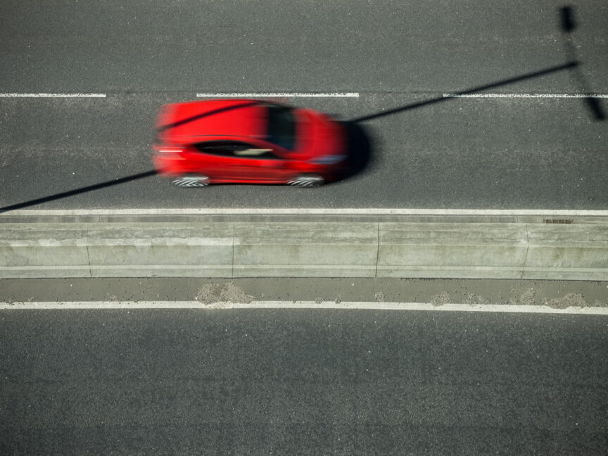 A red car, seen from above, is blurred as it drives along a dual carriageway. Its shape is split by the shadow of an unseen lighting pole which is cast diagonally across the road.