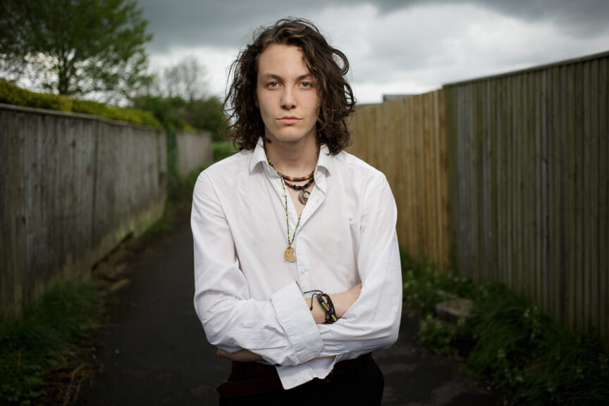 Joe Gander is lit dramatically, standing looking to camera with an alleyway leading off behind him.