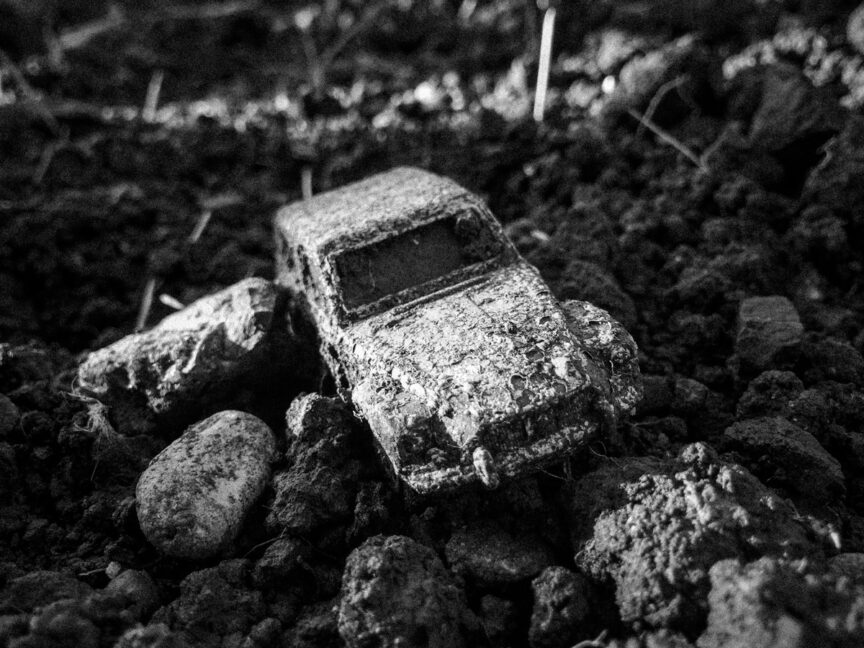 A toy car (a 2CV I think) lies rotting in garden soil.