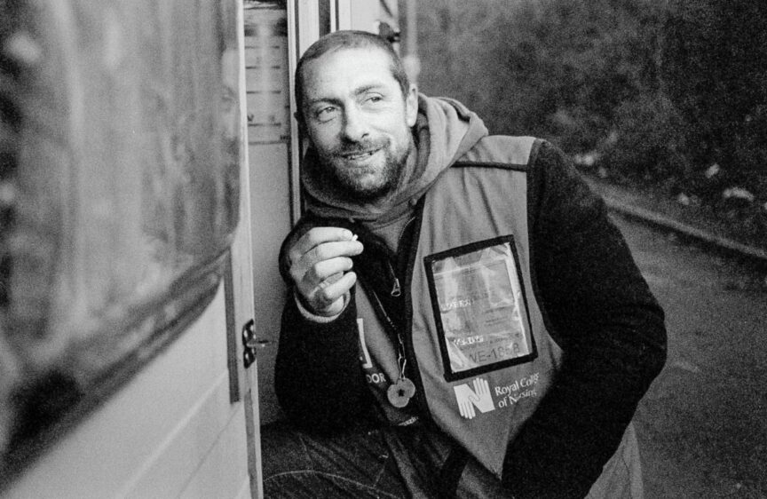 A homeless man leans on the step of his caravan looking away from the camera and smiling as he smokes a cigarette.