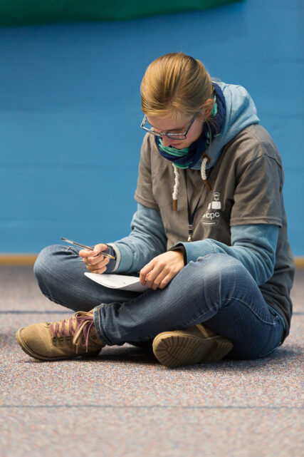 A female student sits alone, cross-legged on the floor with pen and paper.