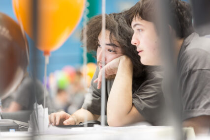 Two male students, seen side-on through balloon strings as they work at their computer.