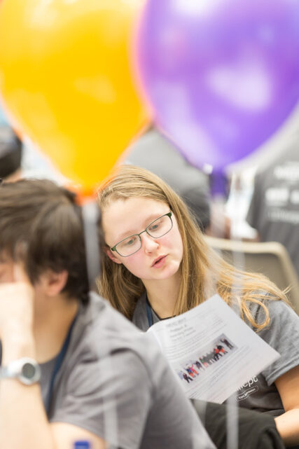 A female student checks over the printed challenge notes, surrounded by balloons and other students which are out of focus.