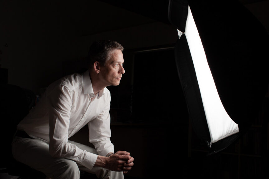 Photographer Tim Gander sits looking into a studio flash softbox which lights his face in an otherwise dark room.