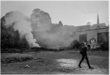 Black and white view of a youth striding off to the right of an open area while to the left in the background a bonfire burns, a thick column of smoke rising into the late afternoon light.