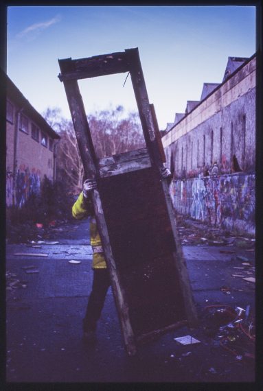 Colour photo of a workman carring a broken door during clean-up. Only his legs, arm and hands are visible as the door obscures the view of the man.