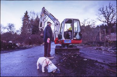 Colour photo of a dog walker chatting to a digger operator in charge of clearing up the site. The dog is wearing a neck cone.