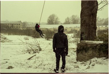 Colour photo of two boys playing in snow. One stands, back to camera, while the other swings away on a rope swing suspended from a tree.