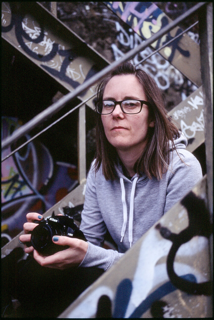 A woman sits on a metal staircase framed by its graffiti-covered rails, holding a camera, wearing glasses and a grey sweat top.