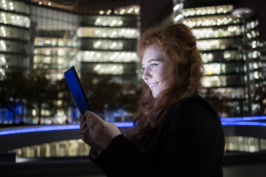 Side-portrait of a young business woman at night in London, her face illuminated by the iPad she is holding up to read. City office block lights illuminate the background.